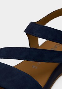Πέδιλο από σουέντ σε ναυτικό μπλε με δύο ευρείες λουρίδες και επενδυμένο καφέ πέλμα. Κομψός σχεδιασμός με επίπεδη σόλα και διακριτικές λεπτομέρειες ραφής.