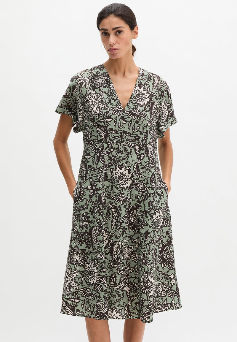 Femme portant une robe verte mi-longue avec des motifs floraux noirs et blancs, manches courtes, col en V, les mains dans les poches latérales.