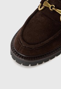 Mocassins en suède marron avec un bout rond, présentant un détail en métal doré et une semelle en caoutchouc noir texturé.