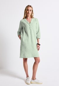 Robe en lin vert clair avec un décolleté en V, des manches 3/4 et une coupe décontractée, présentant une texture subtile et une longueur mi-cuisse. Portée avec des baskets blanches.