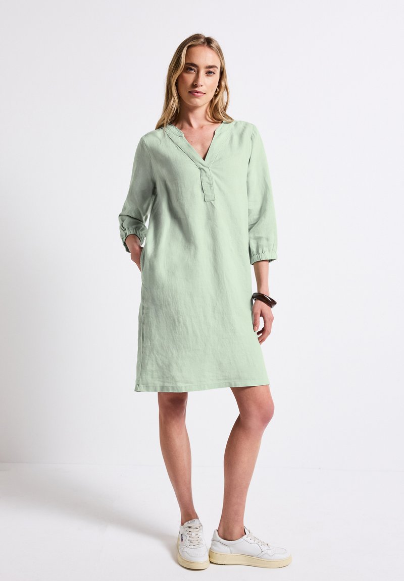 Robe en lin vert clair avec un décolleté en V, des manches 3/4 et une coupe décontractée, présentant une texture subtile et une longueur mi-cuisse. Portée avec des baskets blanches.