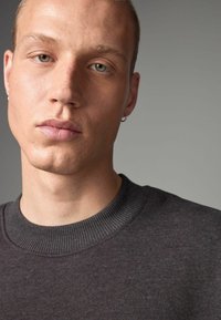Dark gray sweatshirt with a ribbed crew neckline and accented by subtle texture. Close-up of model's face, focusing on features and expression.