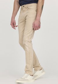 Homme portant un jean slim beige et des baskets blanches, avec des tatouages visibles sur les avant-bras et la main, se tenant contre un fond uni.
