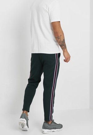 Mężczyzna w czarnych dresowych spodniach z biało-czerwonymi paskami po bokach, białej koszulce i szarych trampkach, stojący tyłem, ukazujący tatuaż na ramieniu.