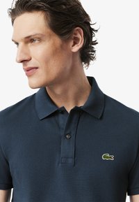 Polo de color azul marino hecho de tela texturizada, con una tapeta de dos botones y un logo de cocodrilo verde bordado en el pecho.