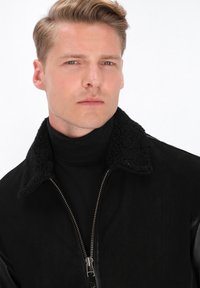 Veste noire en shearling avec col, fermeture éclair, dotée d'un matériau extérieur lisse et d'un col texturé. Le modèle porte un col roulé noir en dessous.
