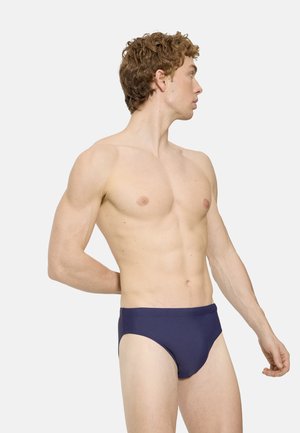 Joven con cabello rizado vistiendo bañador azul marino, de pie sin camiseta y mirando hacia su derecha contra un fondo claro y sencillo.