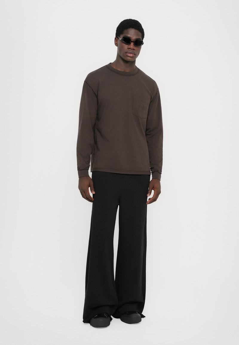 Lange mouw bruin shirt met een borstzak, gecombineerd met wijde zwarte broeken en zwarte schoenen. Model draagt donkere zonnebril.