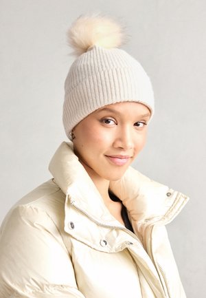 Hvid ribbet strikket hue med en fluffy pompon på toppen, parret med en lys beige puffertjakke med høj krave og metalliske detaljer.