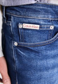 Vaqueros de mezclilla azul oscuro con una etiqueta roja y blanca de "Calvin Klein" en el bolsillo trasero, botones plateados y detalles de costura estándar.