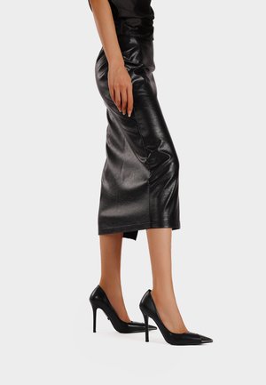 Gonna in pelle nera con silhouette aderente, lunghezza al ginocchio, con una leggera lucentezza. Abbinata a décolleté neri con punta affusolata.