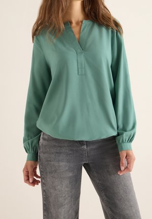 Femme portant un chemisier ample à manches longues de couleur sarcelle avec un col en V et des poignets élastiques, associé à un jean gris délavé.