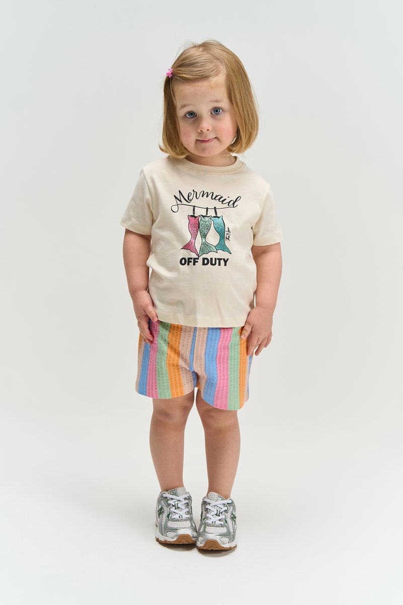Jeune fille blonde debout, portant un t-shirt beige "Mermaid Off Duty", un short rayé pastel, des baskets argentées et une barrette rose.