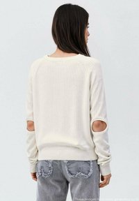 Cremfarbener Strickpullover mit ausgeschnittenen Ärmeln und geripptem Saum, kombiniert mit grauen, ausgewaschenen Jeans mit herzförmigen Patches.