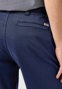 Granatowe spodnie wykonane z gładkiego materiału, z tylną kieszenią z małą metką logo. Styl to luźny krój z szlufkami na pasek.