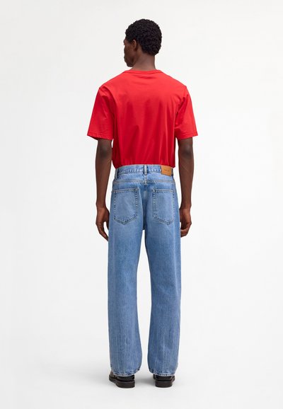 Homme vu de dos portant un t-shirt rouge à manches courtes, un jean bleu clair à jambes droites et des chaussures noires, sur un fond blanc uni.