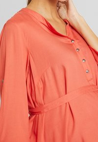 Vestido de camisa coral con escote en V, cierre de botones en el frente y cintura con lazo. Presenta mangas anchas y una tela suave y lisa.