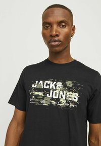 Juodas medvilnės marškinėlis su grafiniu spaudiniu, kuriame rašoma "JACK & JONES" balta spalva, akcentuota abstrakčiu dizainu nuosaikiose spalvose. Trumpomis Rankovėmis.