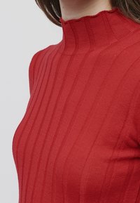 Femme portant un pull col roulé rouge, ajusté et côtelé, présenté du cou jusqu'au milieu du torse sur un fond uni.