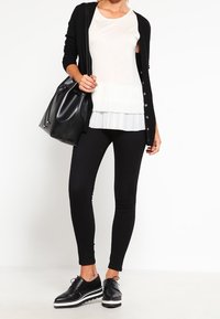 Černý dlouhý svetr přes bílý top s sklady, spárovaný s černými legínami a černými botami s bílými podrážkami. Obsahuje černou kabelku.