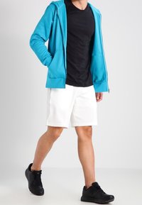 Felpa con zip turchese, t-shirt nera a girocollo, pantaloni corti bianchi e sneakers nere. La felpa ha una texture liscia e tasche frontali.