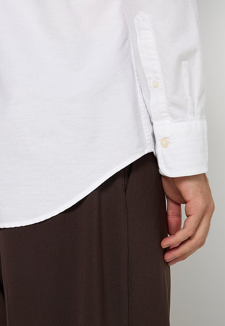 Weißes Langarmhemd mit Knopfmanschetten, aus strukturiertem Stoff und mit abgerundetem Saum, über braunen Hosen liegend.