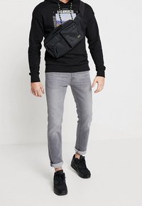 Svart nylon crossbody-väska med flera fickor, bärs över en svart hoodie med grafisk tryck. Gråjeans och svarta sneakers fullbordar looken.