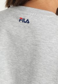 Gray sweatshirt featuring a ribbed neckline and a small red and navy FILA logo embroidered on the back near the neck. Soft cotton material.
