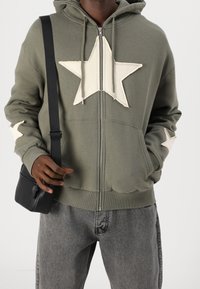Felpa verde con zip caratterizzata da una grande stella bianca sul davanti e piccole stelle sulle maniche; abbinata a jeans grigi e a una borsa a tracolla nera.
