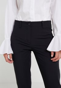 Černé kalhoty na míru s hladkým povrchem a jedním předním knoflíkem, spárované s bílou blůzou s širokými rozšířenými rukávy.