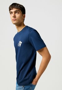 Camiseta de algodón azul marino con cuello redondo, mangas cortas y un gráfico impreso de una cara con un sombrero de vaquero en el pecho.