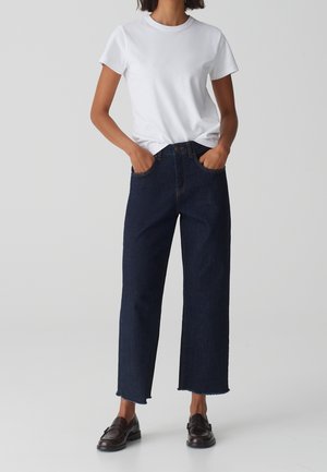 Femme portant un t-shirt blanc à manches courtes, un jean large bleu foncé raccourci avec des ourlets bruts et des mocassins marron foncé, debout les mains dans les poches.