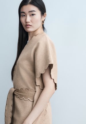 Young woman with long dark hair wears beige scalloped-edge dress with matching belt, gold hoop earrings, against a plain light background.