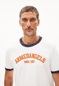 Witte katoenen T-shirt met een marineblauwe ringerkraag en manchetten. Voorzien van oranje geprinte tekst "ARMEDANGELS SINCE 2007" in een vet, gebogen stijl.