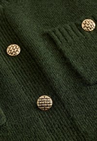 Tummanvihreä neulecardigan, jossa on kultaiset pyöreät napit, kudottu kuvio ja teksturoitu kangas. Cardiganissa on ribbikuviointi ja etutasku.