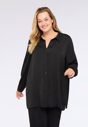 Donna sorridente con lunghi capelli biondi che indossa una camicia nera sbottonata e pantaloni neri, in piedi davanti a uno sfondo chiaro uniforme.