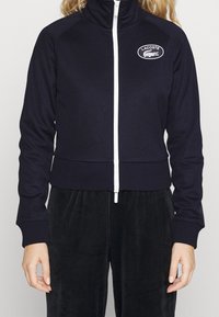 Námořnická zipová bunda s vysokým límcem, bílým zipem a kulatým logem Lacoste. Přiléhavé manžety a pas z žebrované látky.