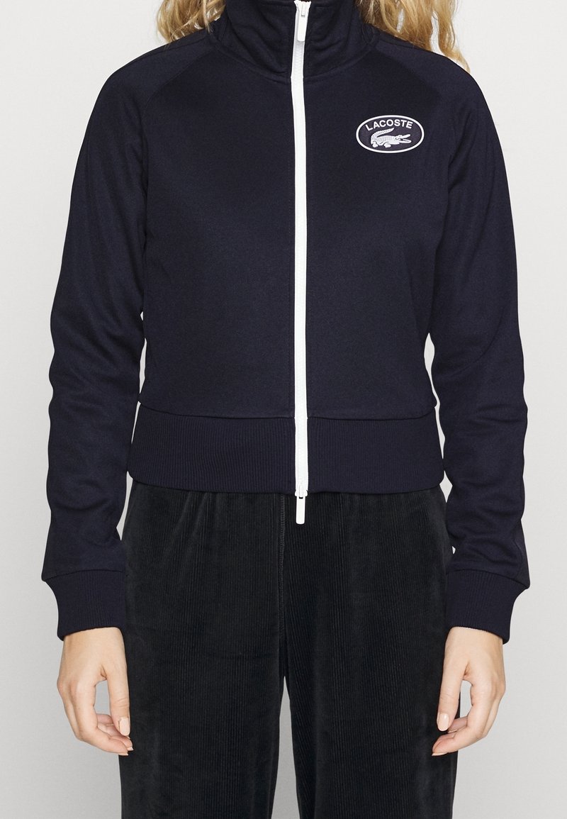Námořnická zipová bunda s vysokým límcem, bílým zipem a kulatým logem Lacoste. Přiléhavé manžety a pas z žebrované látky.