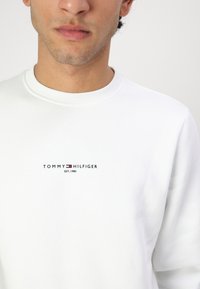 Biely sveter vyrobený z mäkkej tkaniny má dizajn s okrúhlym výstrihom. V strede sa nachádza logo s červeným a modrým textom, ktoré znie: "TOMMY HILFIGER EST. 1985."