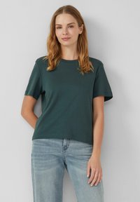T-shirt à manches courtes vert foncé avec un col rond, en tissu de coton doux, coupe décontractée, associé à un jeans en denim bleu clair.
