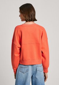 Felpa corallo con vestibilità morbida, colletto rotondo, spalle scese e polsini a coste; abbinata a jeans in denim azzurro chiaro con tasche posteriori.