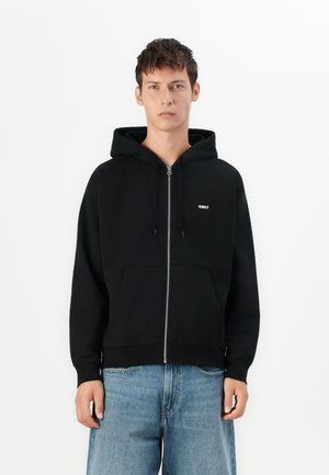 Schwarze Zip-Hoodie aus weichem Stoff, mit einer Fronttasche, Kapuze mit Kordelzug und kleinem weißen "OBEY"-Logo auf der linken Brust.