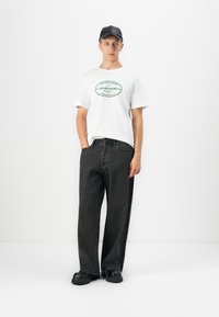 En ung mann som har på seg en hvit grafisk T-skjorte, løse svarte jeans, klumpete svarte sko og en svart baseballcap, står mot en enkel hvit bakgrunn.