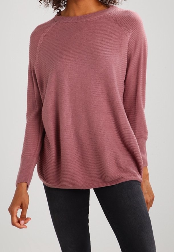 Femme portant un pull en maille côtelée mauve à manches longues raglan et un pantalon noir, debout devant un fond clair uni.