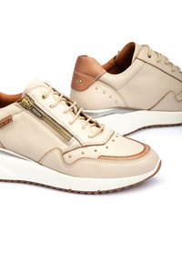 Zapatillas de cuero beige con acentos marrones, detalle de cremallera metálica y adornos de tachuelas. Presentan una suela texturizada y una plantilla acolchada.