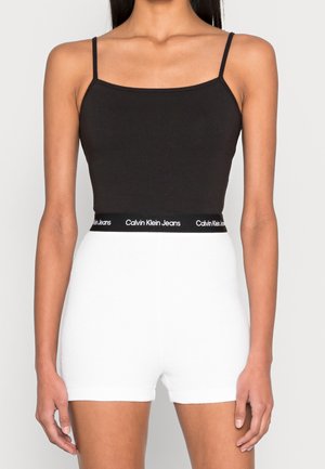 Mujer que lleva una camisola negra ajustada con tirantes finos y pantalones cortos blancos de cintura alta con una cinturilla negra de Calvin Klein Jeans.