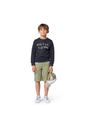 Ragazzo che indossa una felpa nera con testo bianco, pantaloni corti verde oliva e sneaker bianche, che tiene in mano un cappellino beige e verde.