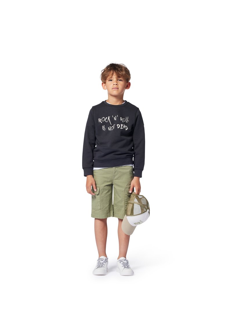 Ragazzo che indossa una felpa nera con testo bianco, pantaloni corti verde oliva e sneaker bianche, che tiene in mano un cappellino beige e verde.