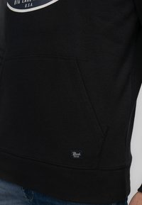 Sudadera negra de tejido de mezcla de algodón, con un bolsillo lateral, puños acanalados y una pequeña etiqueta de marca azul en el dobladillo.