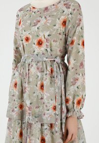 Blomsterkjole i gennemsigtig stof med grønne, pink og orange blomster. Lange ærmer, rund halsudskæring og knyttet taljedetalje med en flagrende nederdel.